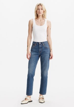 Jeans a vita alta in denim blu, taglio dritto, con cinque tasche, chiusura a bottone e zip, abbinati a un top bianco senza maniche e scarpe.
