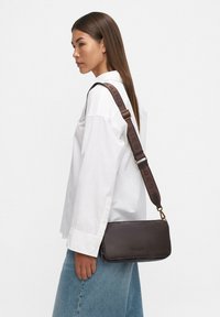 Donna in camicia bianca e jeans blu porta una borsa a tracolla in pelle scura con una larga tracolla di marca.