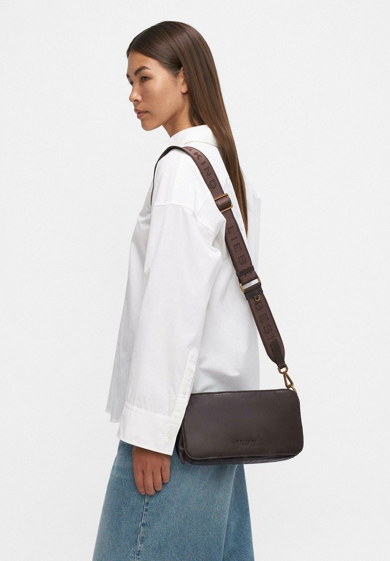 Donna in camicia bianca e jeans blu porta una borsa a tracolla in pelle scura con una larga tracolla di marca.