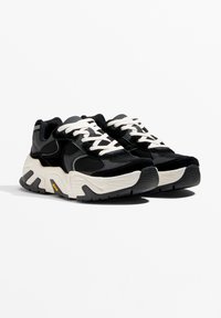 Fekete sportcipők hálós és velúr felsőrésszel, fehér fűzőkkel, valamint egy masszív bézs talppal, fekete díszítésekkel és markáns talpmintázattal.