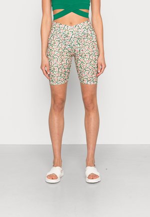Femme portant un short de vélo taille haute à fleurs, un crop top vert et des sandales blanches à bout ouvert, debout devant un fond uni.