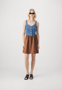 Denim brezrokavnik s sprednjim gumbom, v kombinaciji s svetlo rjavim A-linijskim krilom z vrvico za zategovanje. Obut s črnimi odprtimi sandalmi.