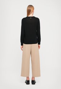 Zwarte longsleeve trui met een ronde hals; gecombineerd met beige ruime broek en zwarte schoenen. Opvallende kleine gouden accent op de trui.