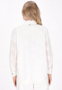 Weißes Langarmhemd aus leichtem Stoff mit floralem Stickmuster, lässiger Schnitt und geknöpften Manschetten. Besitzt einen subtilen Glanz und transparente Ärmel.