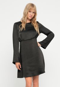 Svart, satin mini kjole med lange bell-ermet og innsnevret liv. Glatt tekstur med rund hals og en lett A-linjeform.