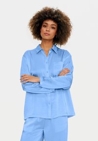 Camicetta azzurro chiaro a maniche lunghe con colletto, tasca frontale e chiusura con bottoni. Texture morbida e vestibilità ampia, abbinata a pantaloni coordinati.