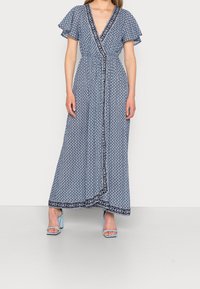 Maxiklänning i blått med vitt geometriskt mönster, v-ringad hals, fjäderlätta ärmar, dragsko i midjan och marinblå detaljer längs kanterna.