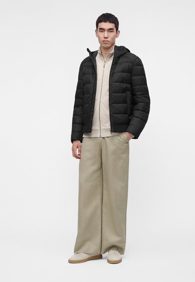 Homme portant une doudoune noire par-dessus un pull beige à fermeture éclair, un pantalon large beige et des baskets blanches, debout avec une main dans la poche.