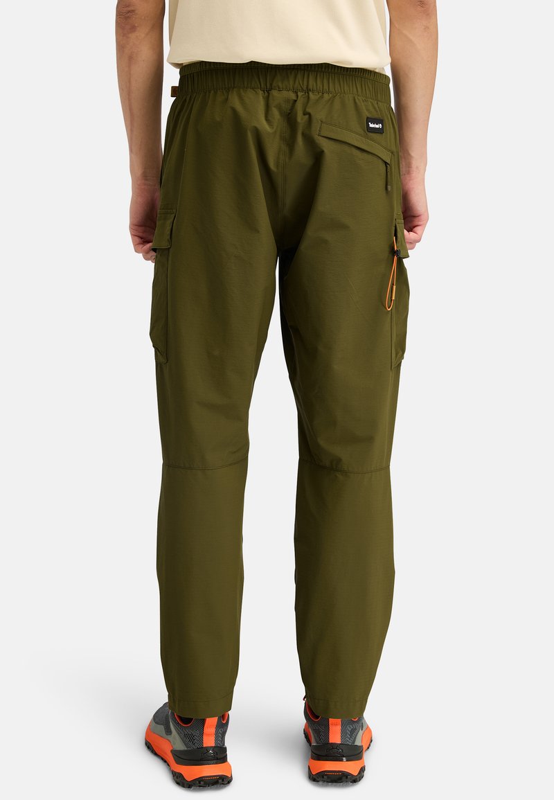 Olivgrüne Cargo-Hosen mit elastischem Bund, Seitentaschen und verstärkten Nähten; kombiniert mit schwarz-orangefarbenen Wanderschuhen.