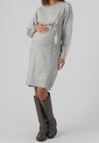 Grå stickad gravidklänning med långa ärmar och ett bälte i midjan. Modellen har på sig höga bruna läderstövlar. Slät textur, knälång design.