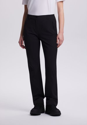 Person iført sorte bukser med lige ben og lynlåslommer i siden, sorte sko og en hvid kortærmet skjorte, stående på en ensfarvet baggrund.