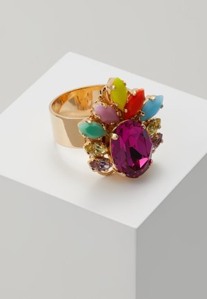 Bague en or avec une grande pierre ovale violette entourée de petites pierres multicolores de différentes formes, présentée sur un bloc d'exposition blanc.