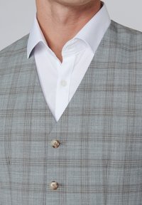 Pilkas langelių rašto liemenė, pagaminta iš tekstūruotos medžiagos, su V formos dekolete ir dviem mygtukais, derinama su balta marškinėliais su apykakle.