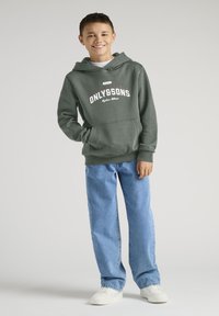Zielona bluza z kapturem z białym napisem "ONLY&SONS", w połączeniu z jasnoniebieskimi dżinsami i białymi trampkami. Model stoi swobodnie, z jedną ręką w kieszeni.