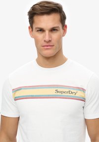 Camiseta de algodón blanca con un diseño de rayas horizontales en azul, amarillo y rojo, con el logotipo "SuperDry" en negro en el pecho.
