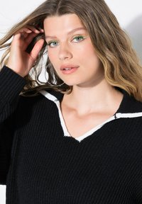 Jonge vrouw met los golvend haar, draagt groene eyeliner, grote ringoorbellen en een zwart gebreid sweater met een witte kraagdetail.