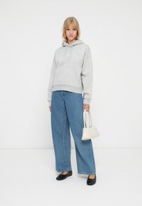 Donna che indossa un hoodie grigio, jeans a gamba larga blu, ballerine nere, mentre tiene una piccola borsa bianca su uno sfondo bianco semplice.