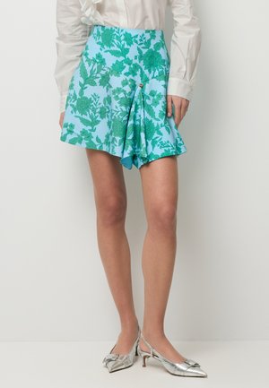 Lichtblauwe rok met bloemenprint en een plooidetail met knopen aan de voorkant, gedragen met zilveren kittenheels en een witte blouse.