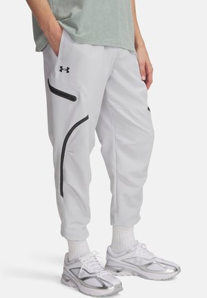 Mann trägt hellgraue, schmal zulaufende Hose mit schwarzen gebogenen Streifen, weiße Socken und weiße Sportschuhe, steht mit einer Hand in der Tasche.