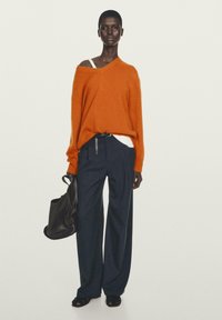 Übergroßer orangefarbener Pullover, weißes Top darunter, marineblaue, gestreifte, weit geschnittene Hosen, schwarze Ledertasche und schwarze Sandalen. Glatte Texturen.