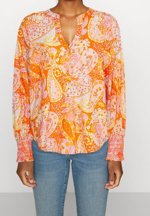 Blouse orange à manches longues avec imprimé cachemire rose et jaune, poignets smockés, portée avec un jean bleu sur une personne debout.