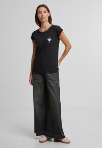 Schwarz kurzärmliges T-Shirt mit einem Diamantlogo auf der Brust, kombiniert mit weit geschnittenen dunkelblauen Jeans und hellen Sneakers. Einfaches, klares Design.