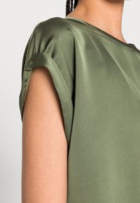 Olivgrüne Bluse mit kurzen hochgekrempelten Ärmeln, glatter Satinstruktur und lockerem Schnitt, der eine dezente Drapierung an den Schultern zeigt.