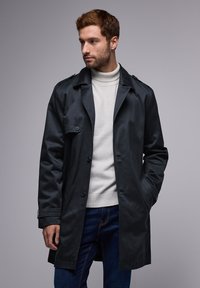 Trench coat de color azul oscuro con cinturón, botones visibles y dos bolsillos delanteros, usado sobre un cuello de tortuga de color claro y jeans azules.