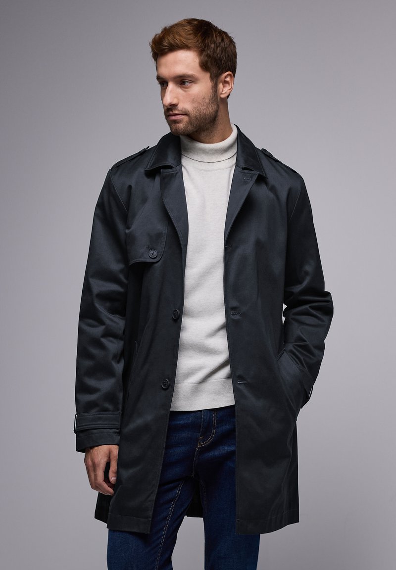 Trench coat de color azul oscuro con cinturón, botones visibles y dos bolsillos delanteros, usado sobre un cuello de tortuga de color claro y jeans azules.