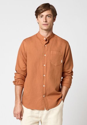 Junger Mann steht mit den Händen in den Taschen, trägt ein rostfarbenes Hemd mit Stehkragen und helle Hosen.