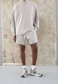 Světle šedá mikina s vysokým límcem a dlouhými rukávy, odpovídající šedé kraťasy a šedé běžecké boty s bílými akcenty. Texturované pozadí.