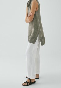 Tunic senza maniche, a costine, di colore verde oliva con colletto alto e spacchi laterali, abbinata a pantaloni larghi bianchi e sandali neri.