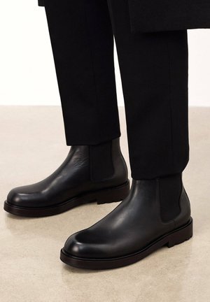 Mustad mustad Chelsea saapad elastsete külgpaneelidega, ümar varvas ja paks tekstureeritud tald. Kantud mustade pükstega heledal pinnal.
