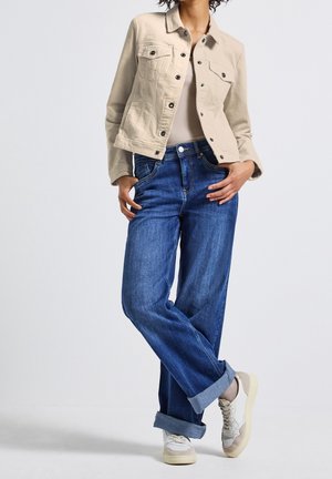 Femme portant une veste en jean beige, un top beige clair, un jean bleu retroussé et des baskets blanches, posant avec une jambe croisée sur l'autre.