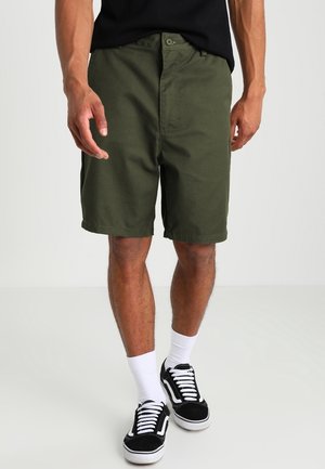 Hombre vistiendo pantalones cortos verde oliva, camiseta negra, calcetines blancos hasta el tobillo y zapatillas negras de caña baja, de pie sobre un fondo claro liso.