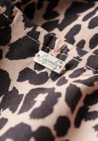 Leopardprint stof in beige en zwart, met een zilveren Superdry logo-label dat is bevestigd met een lichtbruine touw. Geweven oppervlakdetail zichtbaar.