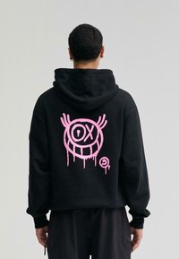 Sudadera con capucha negra que presenta un gráfico en estilo graffiti rosa en la parte posterior, con un rostro estilizado y acentos goteantes. Puños y dobladillo acanalados.