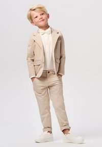Beige pak met een vlinderdas, wit overhemd en beige broek. De jongen staat met zijn handen in zijn zakken, en draagt witte sneakers. Glad textuur van de stof.