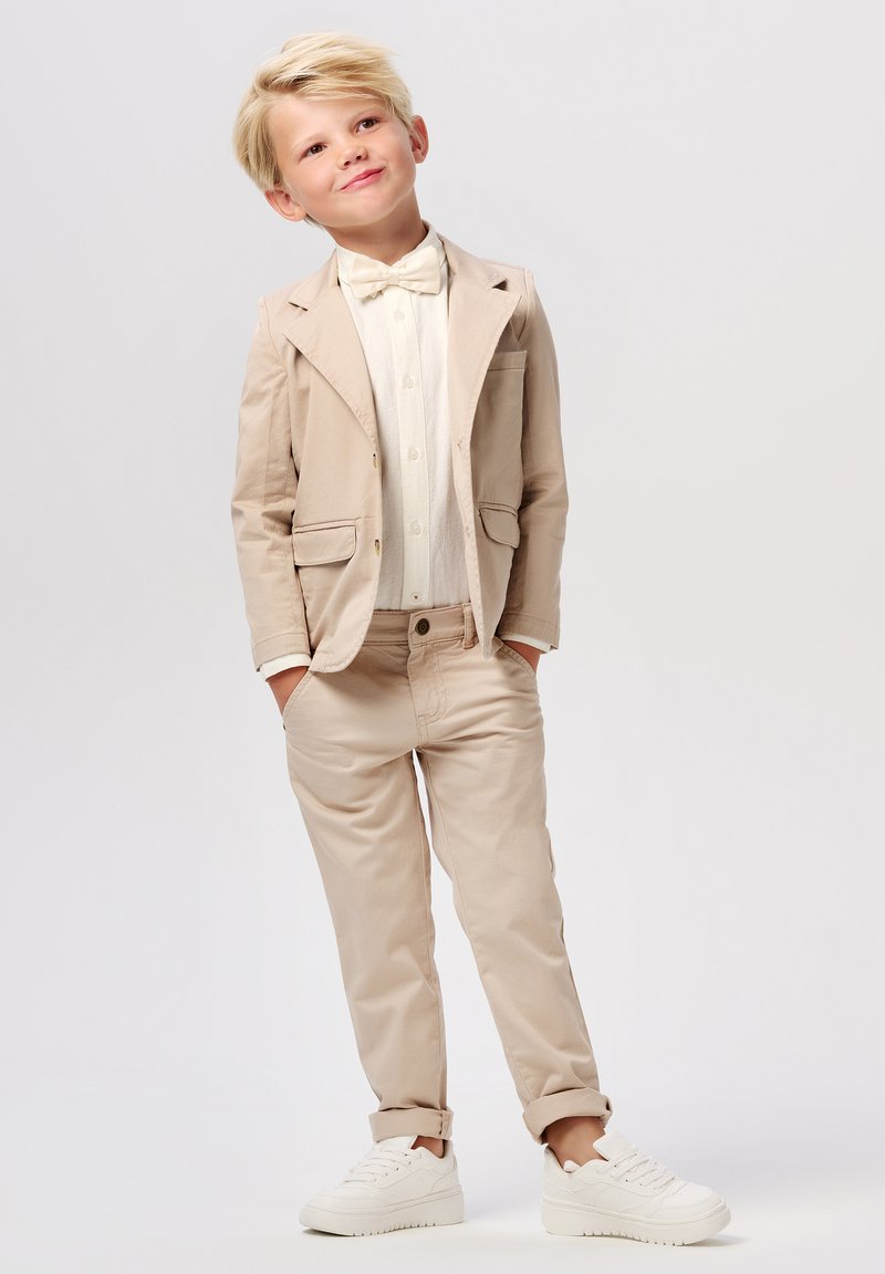 Beige pak met een vlinderdas, wit overhemd en beige broek. De jongen staat met zijn handen in zijn zakken, en draagt witte sneakers. Glad textuur van de stof.
