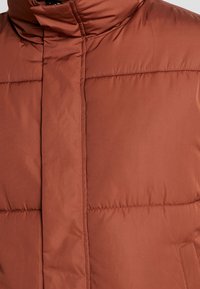 Veste matelassée couleur rouille avec col haut et fermeture éclair dissimulée à l'avant, cousue en sections horizontales.