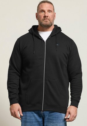 Homme barbu portant un sweat noir à fermeture éclair sur un t-shirt blanc et un jean bleu, debout face à l'avant devant un fond uni et clair.
