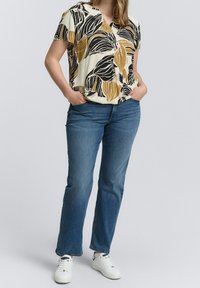 Kortærmet top med sort og sennepsfarvet bladmønster, parret med blå denim flared jeans og hvide sneakers. Teksturen fremstår glat.