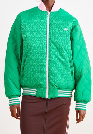 Personne portant une veste matelassée verte avec une fermeture éclair rose, un devant à motifs, des poignets et un ourlet rayés, et une jupe marron avec des bandes latérales.