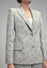 Femme portant un blazer gris et bleu clair à motifs, sur mesure, avec des boutons argentés et un pantalon assorti.