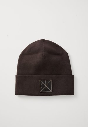 Brown knitted beanie with a folded cuff featuring a small, square logo in contrasting color. Smooth texture and rounded top design.
