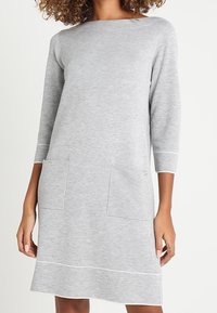 Donna che indossa un vestito di maglia grigio chiaro, lungo fino al ginocchio, con maniche a tre quarti e due tasche frontali a toppa.