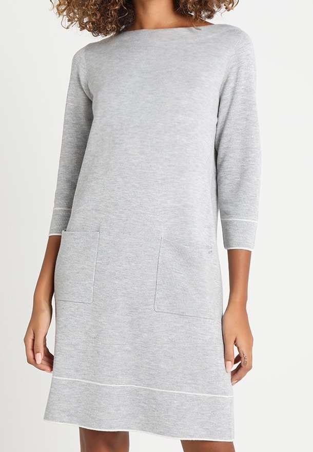 Donna che indossa un vestito di maglia grigio chiaro, lungo fino al ginocchio, con maniche a tre quarti e due tasche frontali a toppa.