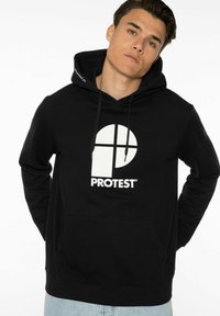 Felpa nera con cappuccio realizzata in un tessuto morbido, caratterizzata da un grande logo circolare bianco con un design a croce e testo. Include una tasca frontale e cordini per il cappuccio.