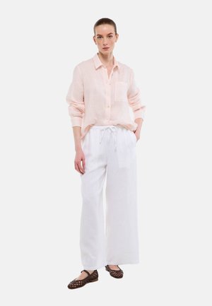 Femme debout portant une chemise rose clair à boutons, un pantalon blanc à jambes larges et des chaussures plates tressées foncées sur un fond blanc.