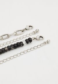 Chaîne en ton argenté avec des sections alternées : maillons lisses, perles texturées noires et grises, et une perle transparente. Comprend plusieurs fermoirs.
